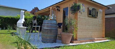 A small wooden cabin with a porch, a barbecue grill, and a dartboard.
