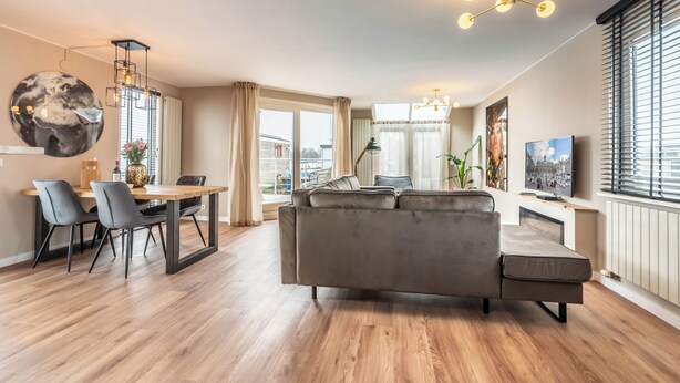 A modern living room with a grey sofa, wooden flooring, a dining area with chairs, a chandelier, and a TV. - Spacious 120m2 Watervilla With Sauna Near Amsterdam And Schiphol. (Aalsmeer)