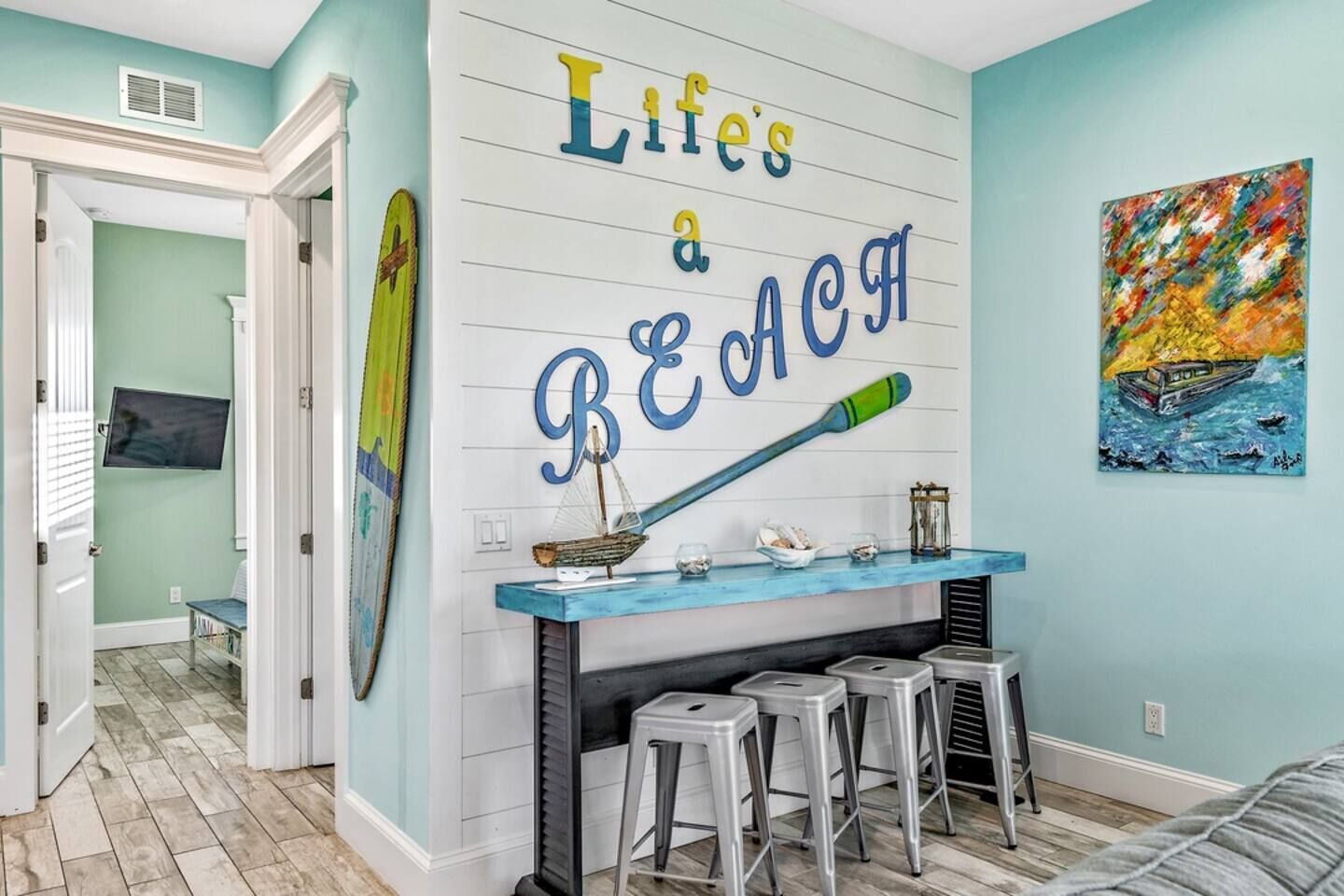 A room with a wall sign reading "Life's a BEACH," a small bar area with stools, a surfboard decoration, and a colorful painting of a boat.