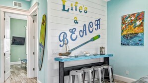 Et værelse med et skilt på væggen, der siger "Life's a BEACH", et lille barområde med barstole, en surfbræt-dekoration og et farverigt maleri af en båd.