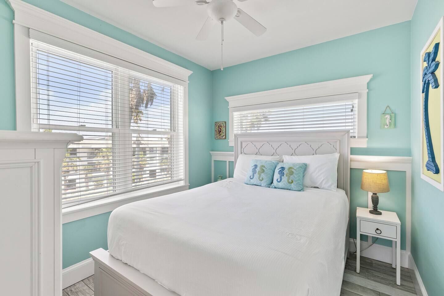 A bedroom with a bed, two windows with blinds, a nightstand with a lamp, and a wall with a framed picture.