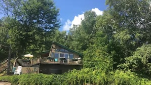 A cabin with a deck, surrounded by dense forest and a body of water.