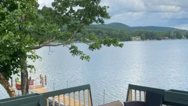 A deck with wicker chairs and a glass table overlooking a lake with a dock and greenery in the background.