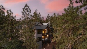 A two-story cabin surrounded by dense forest, with a visible deck and a large window on the upper floor.