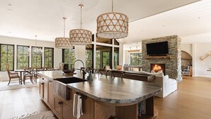 A spacious kitchen with a large island, pendant lights, a stone fireplace, and a TV mounted on the wall.
