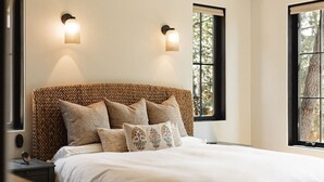 A bedroom with a woven headboard, a bed with white bedding, two wall-mounted lights, and a window with a view of trees.