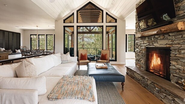 A spacious living room with a stone fireplace, large windows, and a high ceiling.