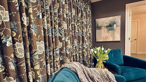 Una sala de estar con sofás color turquesa, una cortina decorativa con motivos florales y un jarrón con flores blancas.