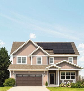 A two-story house with a garage, a front porch with chairs, and solar panels on the roof.