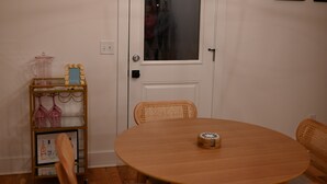 A round wooden table with a small decorative item in the center, a wicker chair, and a metal shelf with glassware and a coffee maker to the left. - Nashville Rodeo House (Nashville)