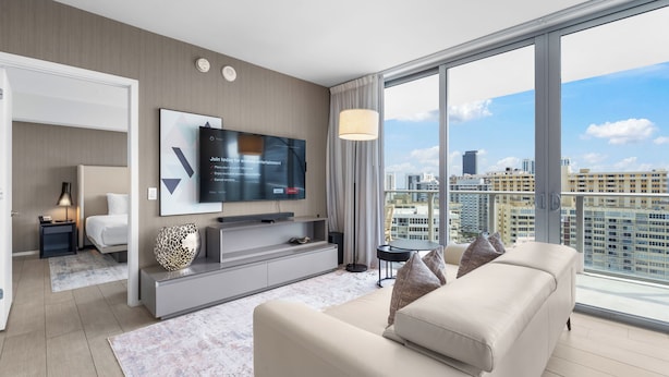 A modern hotel room with a large window offering a city view, a flat-screen TV mounted on the wall, a sofa, and a small side table with a lamp.