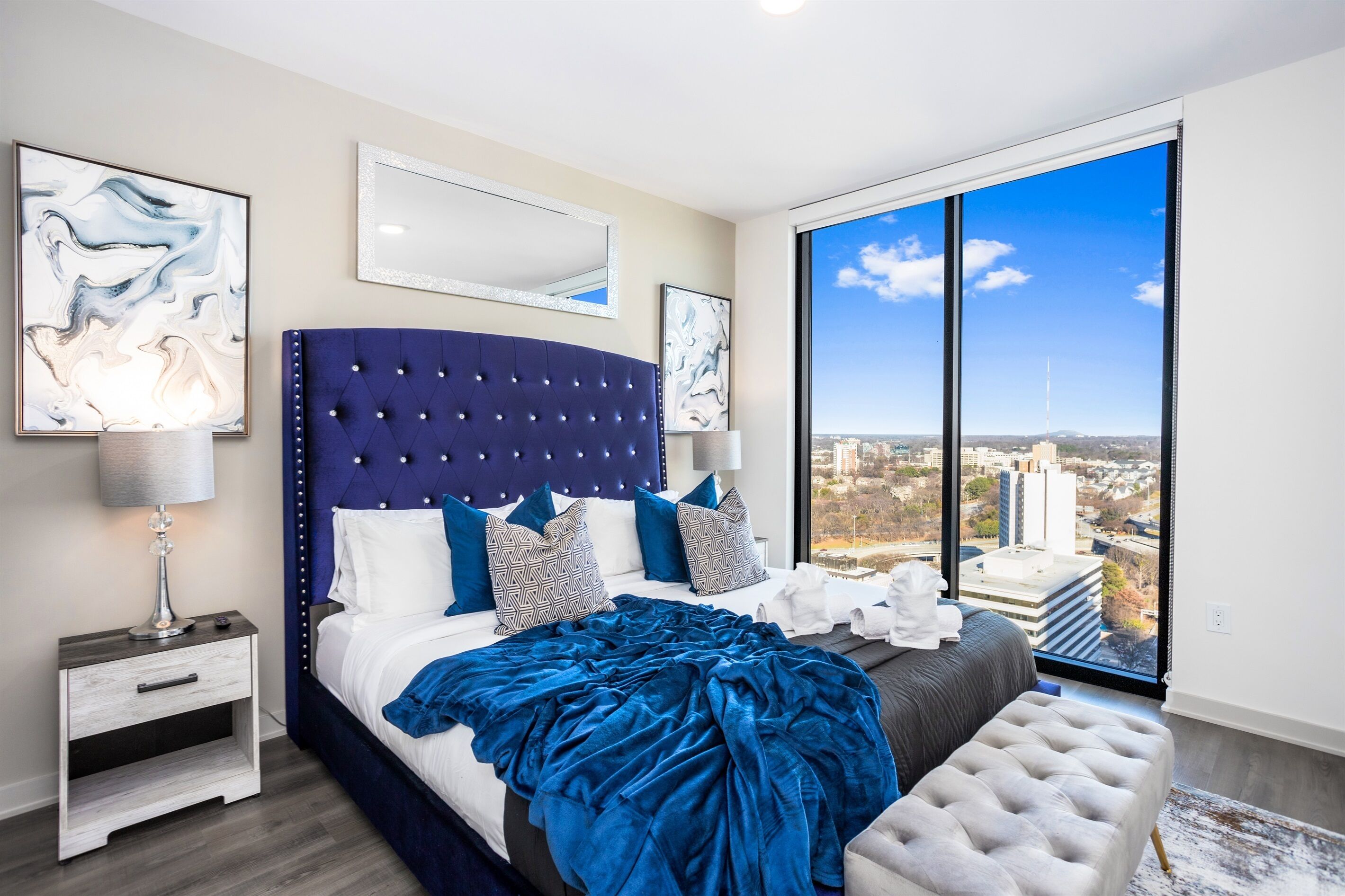 A modern bedroom with a large bed, a nightstand, a dresser, and a large window with a city view.