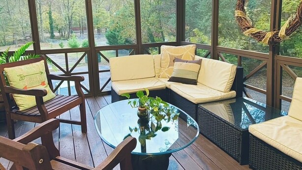 A covered outdoor seating area with wicker furniture and a glass table, overlooking a wooded area. - Black Bear's Riverside Chalet (Helen)