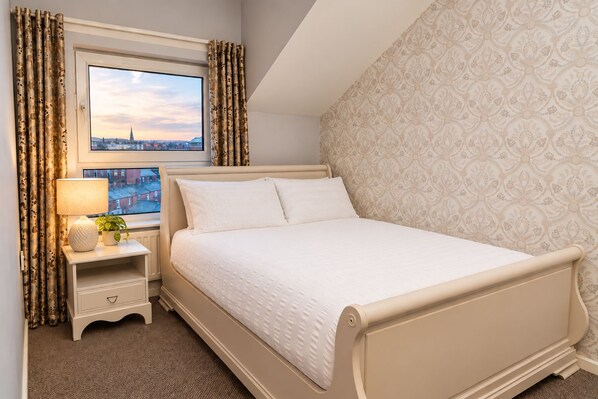 A bedroom with a bed, a bedside table with a lamp and a plant, a window with curtains, and a patterned wall.