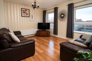 A living room with two leather sofas, a television on a wooden stand, a wall-mounted sunburst mirror, and a window with curtains.