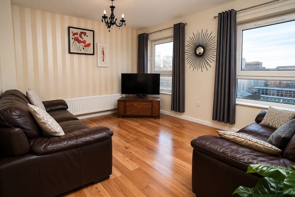 A living room with two leather sofas, a television on a wooden stand, a wall-mounted sunburst mirror, and a window with curtains.
