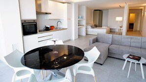 Ein modernes Hotelzimmer mit Küche, Essbereich und Wohnbereich. Es beinhaltet einen runden Esstisch, weiße Stühle, ein Sofa und einen kleinen Beistelltisch mit einer Lampe.