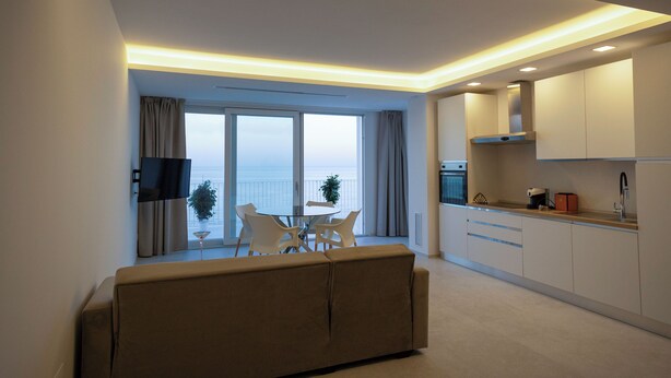 Ein modernes Wohnzimmer mit einer Couch, einem Esstisch und Stühlen, einer Küche mit Schränken und Geräten, sowie einem großen Fenster mit Meerblick.