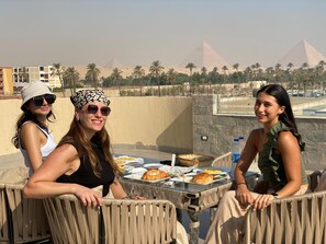 Three people seated at an outdoor table with food, overlooking palm trees and pyramids. - Galleria Pyramids View Hotel (Giza)