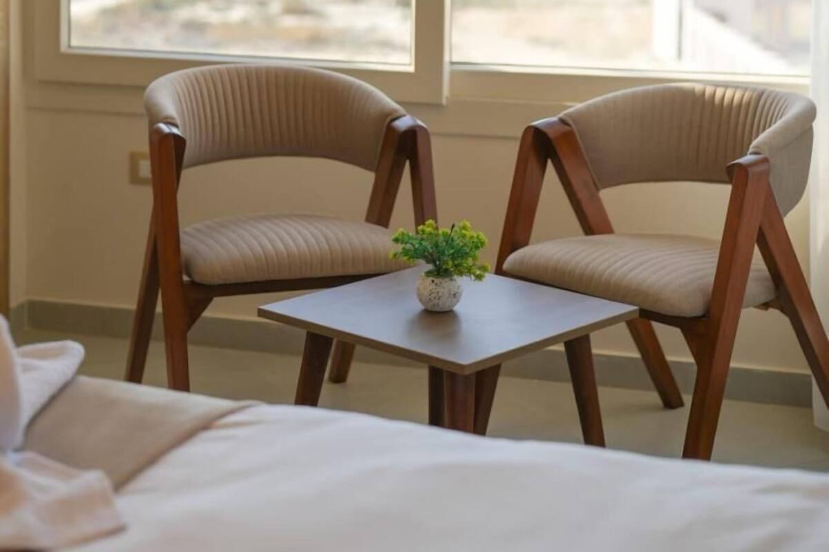 A hotel room with two wooden chairs, a small table with a potted plant, and a bed. A large window offers a view of a pyramid and cityscape.