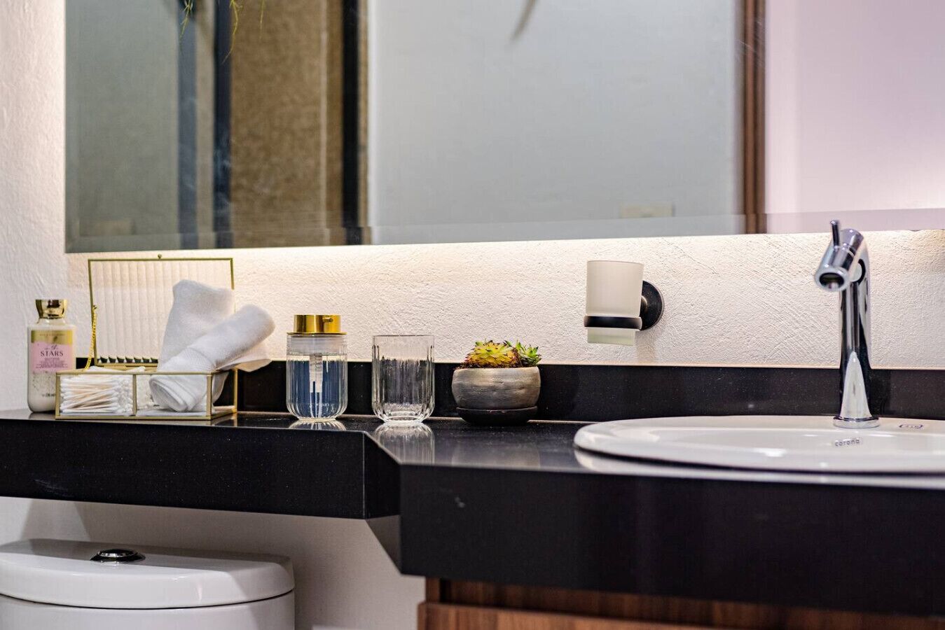 Casa de banho com bancada preta, lavatório branco, espelho, porta-toalhas, uma pequena planta em vaso e um frasco de loção.