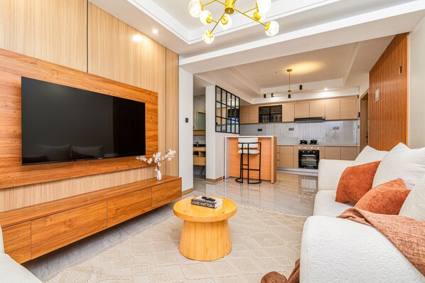 A modern living room with a large flat-screen TV, wooden furniture, a round coffee table, and a sofa with cushions. - Stylish & Modern 1 BR | Pool, Gym & Roof Terrace | Kileleshwa (Nairobi)