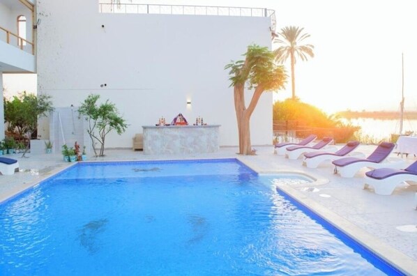 A swimming pool with lounge chairs and a bar area, surrounded by plants and a view of the sunset. - Eyaru Palace (Luxor)