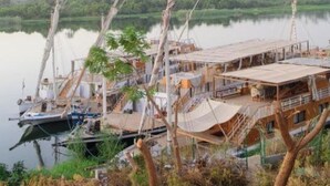 A riverside lodge with a wooden deck and a boat docked nearby. - Eyaru Palace  (Luxor)