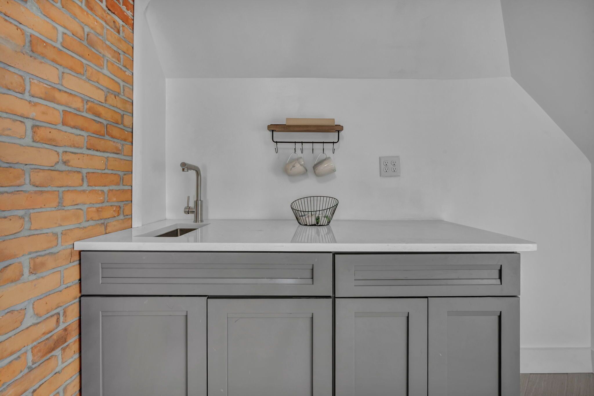 Une cuisine avec des meubles gris, un plan de travail blanc, un porte-ustensiles suspendu et un mur en briques.