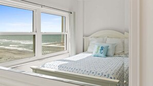 Un dormitorio con un ventanal grande con vistas a la playa, una cama con colcha estampada y cabecera blanca.