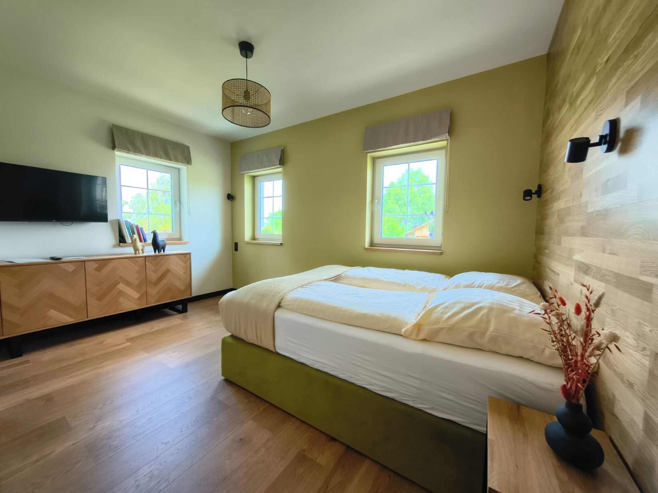 A bedroom with a bed, a TV, a wooden dresser, and two windows with curtains.
