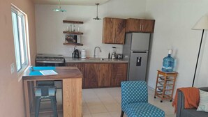 Eine moderne Küche mit Holzausstattung, einem eingebauten Herd und einem Kühlschrank. Es gibt einen Essbereich mit Tisch und Stühlen, sowie einen blauen Stuhl mit Muster. Ein Wasserspender steht auf einem kleinen Tisch neben der Couch.