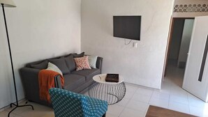 Ein Wohnzimmer mit einer Couch, einem gemusterten Sessel, einem kleinen runden Couchtisch und einem an der Wand montierten Flachbildfernseher.