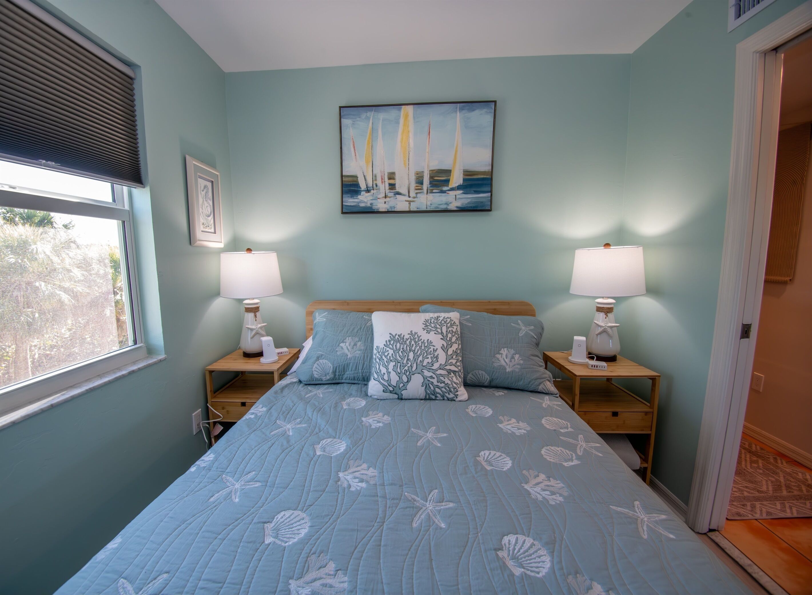 A bedroom with a bed, two bedside tables with lamps, a painting of sailboats, and a window with blinds.