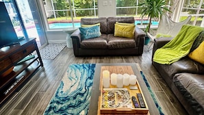 A living room with a brown leather sofa, a coffee table with candles and remote controls, and a large window offering a view of a swimming pool and greenery. - YOUR PRIVATE RETREAT NEAR SIESTA KEY BEACH! (Sarasota)