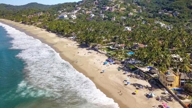Une plage aux eaux turquoise et limpides, un rivage sablonneux et une colline parsemée de bâtiments et de palmiers.
