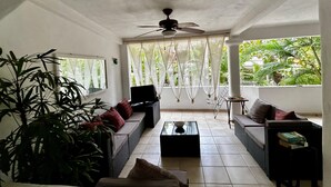 Un vaste espace de vie comprenant deux canapés, une table basse et un ventilateur de plafond. De grandes fenêtres ornées de rideaux légers laissent entrer la lumière naturelle et offrent une vue sur la verdure environnante.