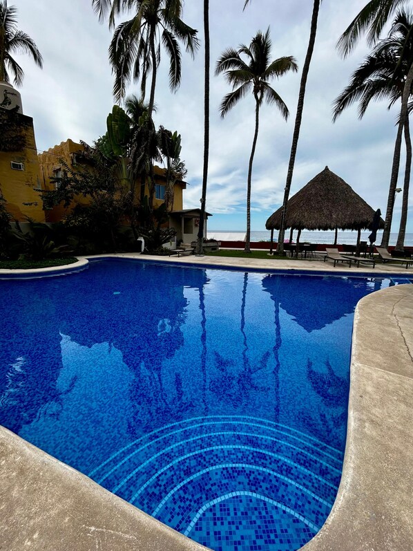 Una piscina azul cristalina rodeada de palmeras y una estructura con techo de paja, con vista al océano al fondo.