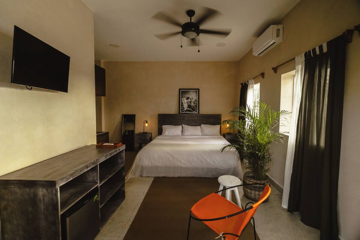 Quarto de hotel com cama, escrivaninha, cadeira, ventoinha no teto, ar condicionado e janela com cortinas.