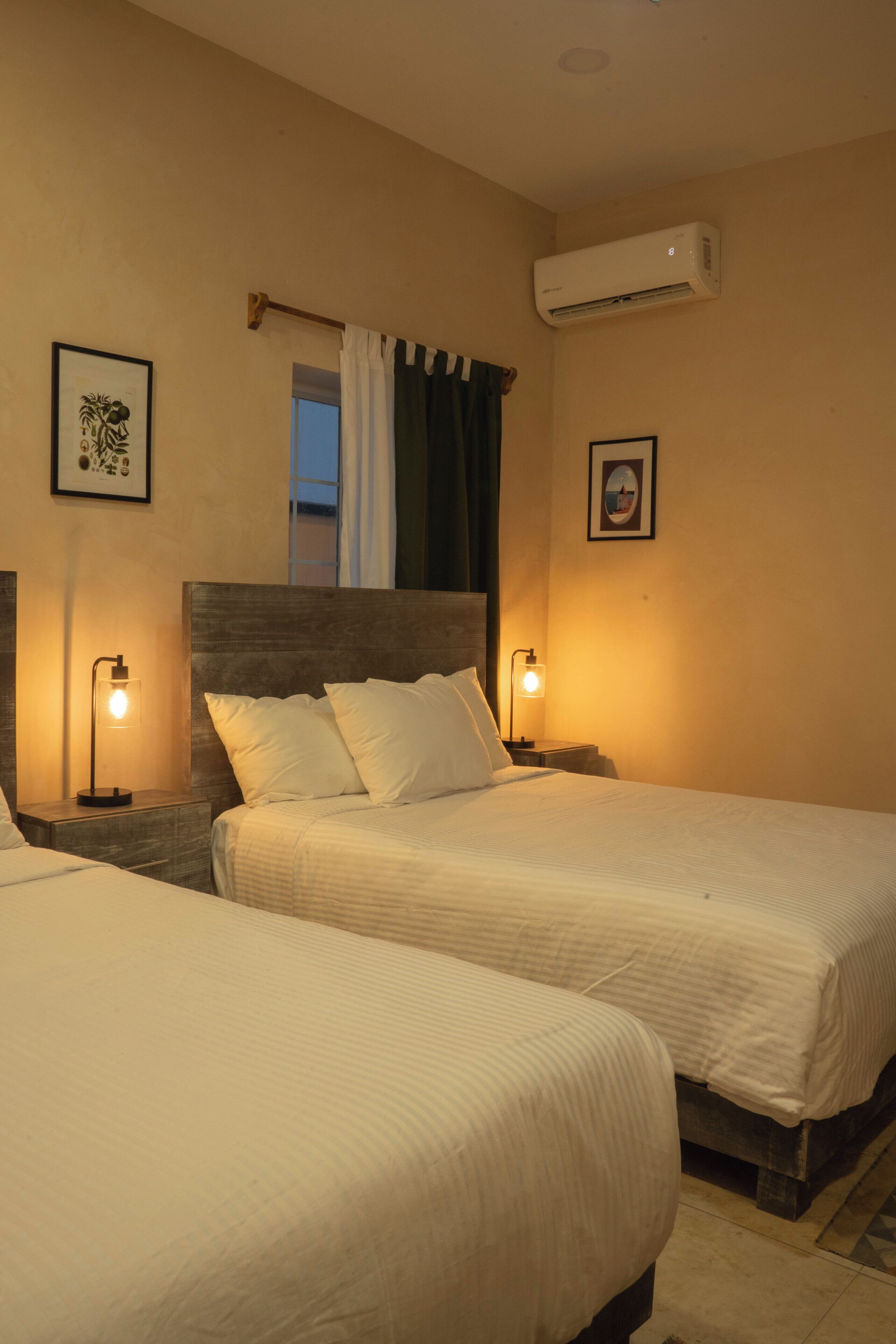 Habitación de hotel con dos camas, una mesita de noche con lámpara, aire acondicionado y cuadros en la pared.