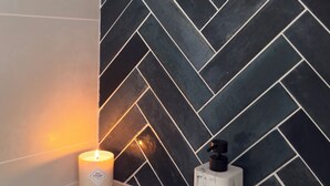 Ein Badezimmer mit schwarzer Fliesenrückwand im Fischgrätmuster, einer brennenden Kerze und einem Marmel-Seifenspender auf einer beige farbenen Arbeitsplatte.