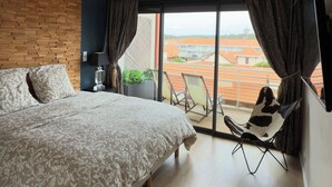 Ein Schlafzimmer mit einem großen Bett, einem Holzkopfende, Blick auf Gebäude durch ein großes Fenster und einem Balkon mit Stühlen.