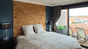 Ein modernes Schlafzimmer mit Holzkopfstütze, einem großen Bett mit gemusterten Betttextilien, einem Nachttisch mit Lampe und Blick auf die Stadt durch ein großes Fenster.