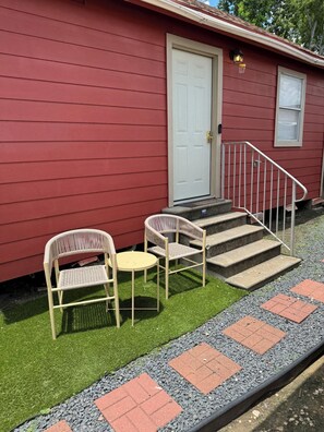 A red building with a white door, a small outdoor seating area with wicker chairs and a table, and a set of stone steps leading to a white door.