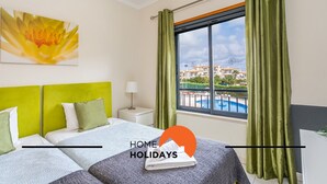 A hotel room with two beds, a window with a view of a swimming pool and buildings, and a green curtain.