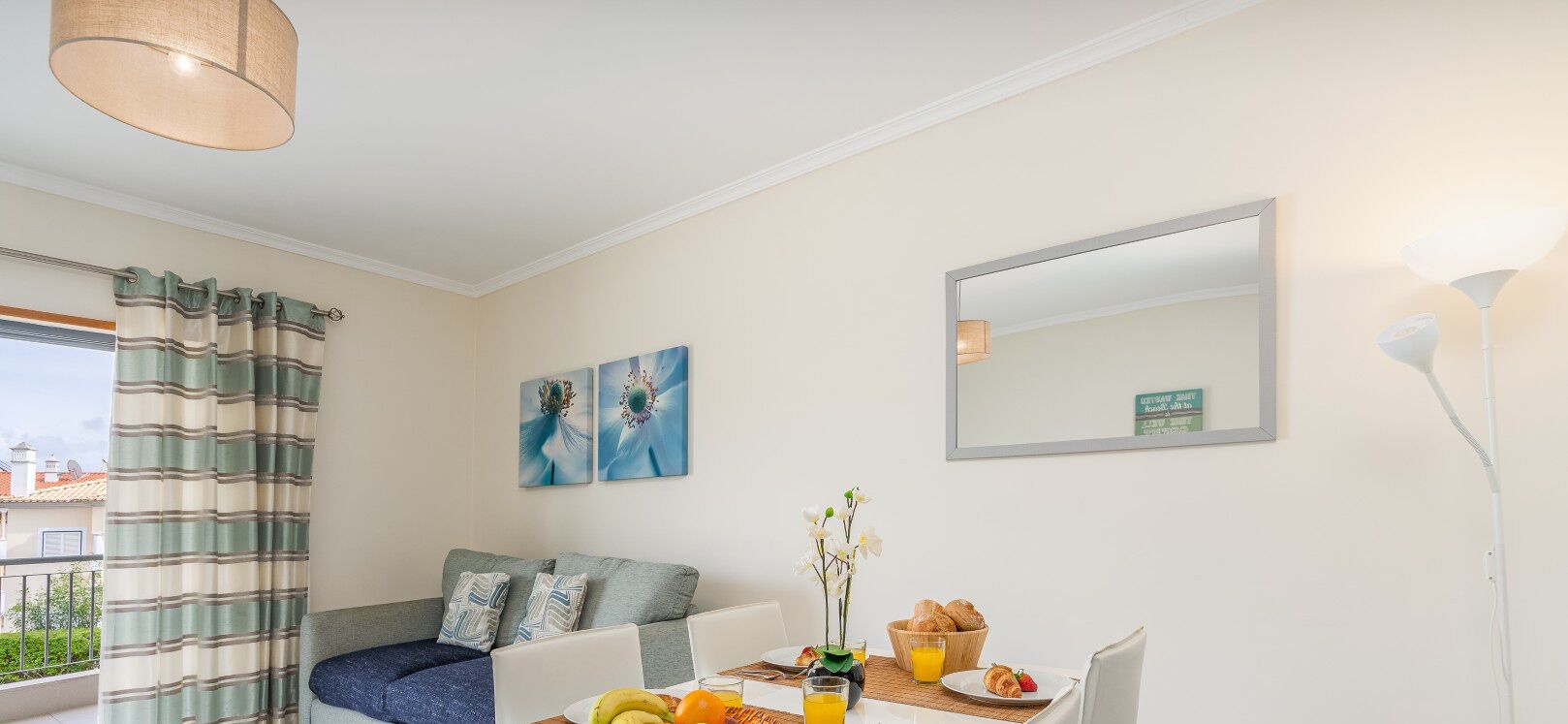 Un salon moderne avec un canapé, une table à manger où sont disposés des aliments pour le petit-déjeuner, un grand miroir et des œuvres d’art accrochées au mur.