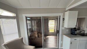 Amplia sala de estar con un sillón beige, una cocina con armarios blancos y un ventanal corredizo que da a un balcón.