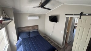 Un dormitorio con cama, ventilador de techo, televisor montado y ventana con persianas.