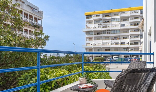 Un balcon agrémenté de chaises en osier et d’une petite table, d’où l’on peut admirer un immeuble doté de balcons et d’une rambarde bleue.