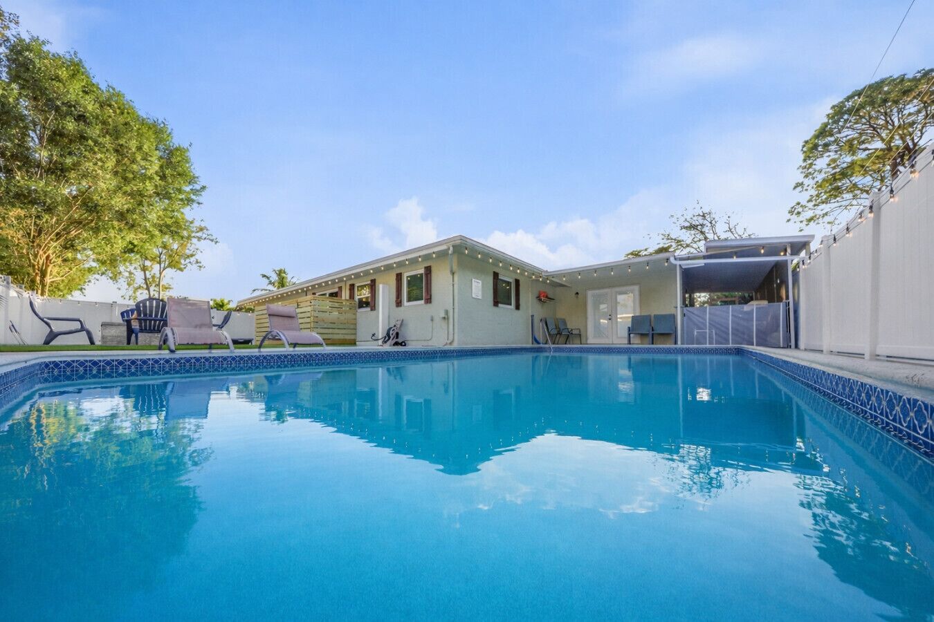 A swimming pool with lounge chairs, a covered patio, and a building with string lights.
